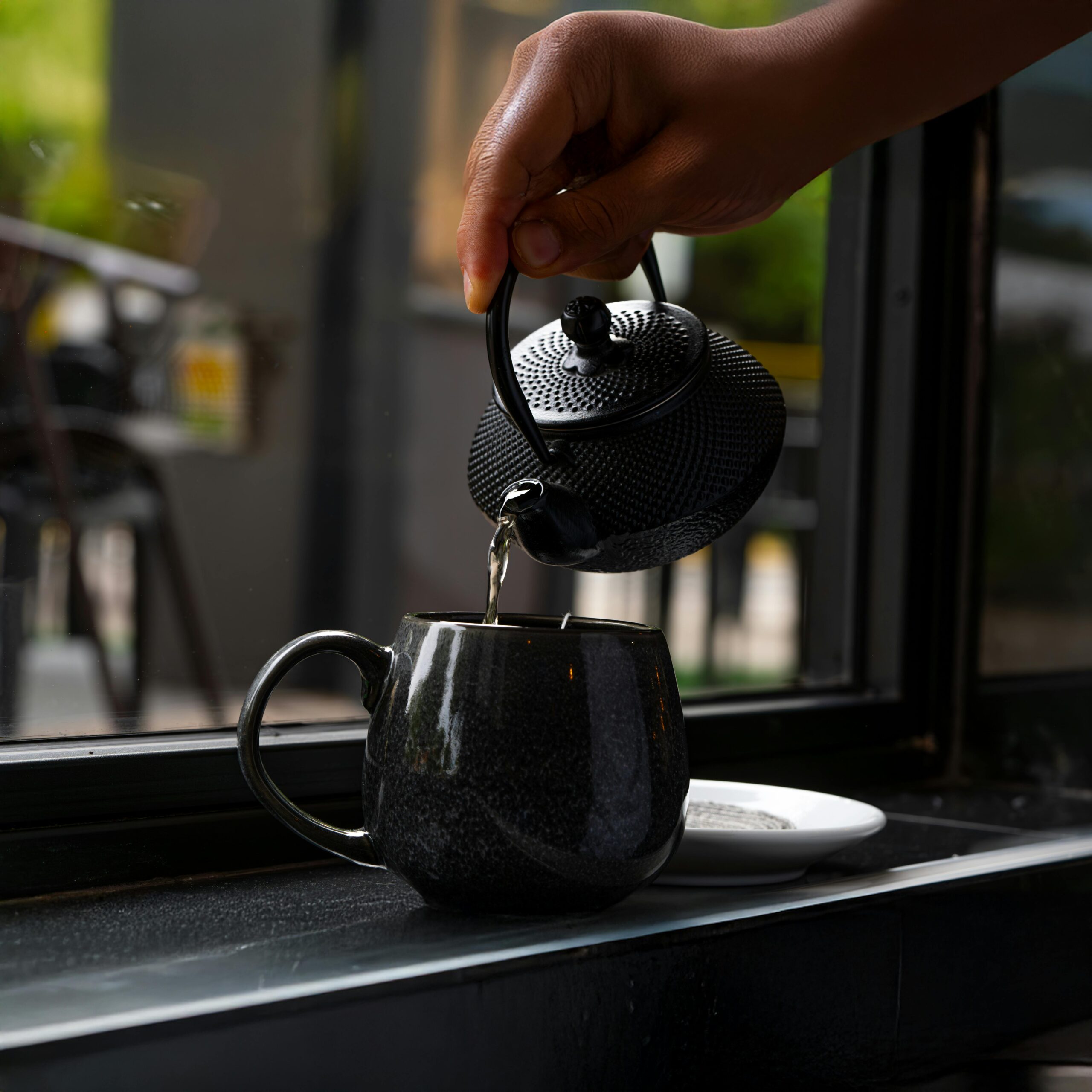 Close-up of hand pouring hot tea from black cast iron teapot into mug indoors.