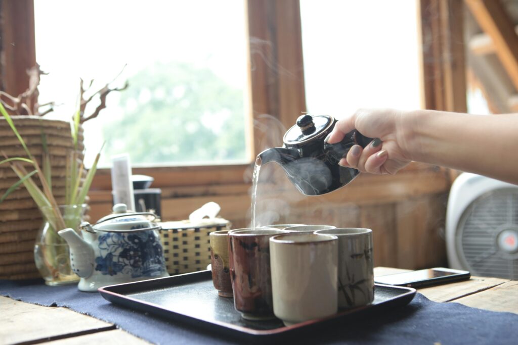 A hand pours steaming hot tea into ceramic cups with a black teapot on a wooden table.