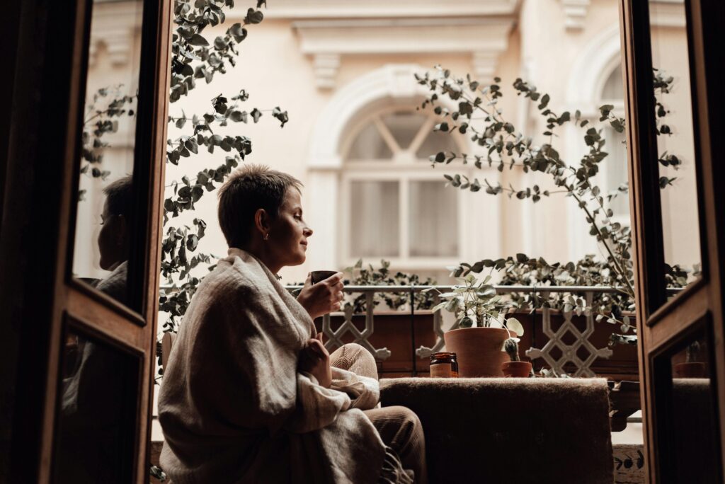 Woman wrapped in a blanket sipping tea on a charming balcony with plants.