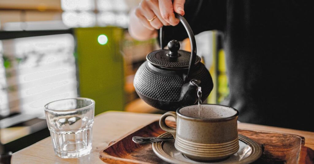 A person pours hot tea from a cast iron teapot into a cup on a wooden tray.
