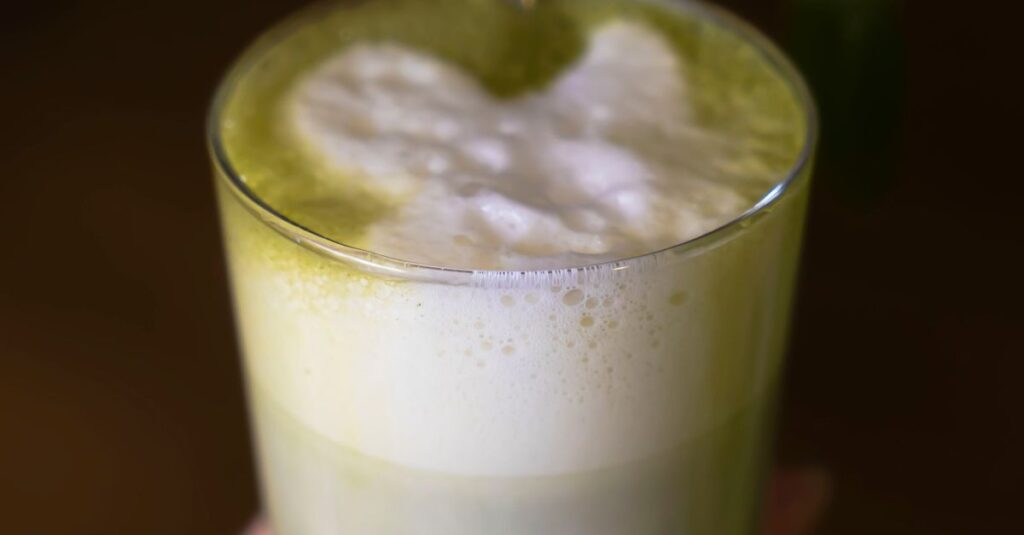Close-up of a hand holding a frothy green matcha latte in a glass.
