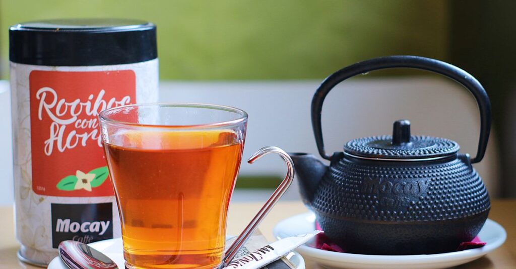 A stylish black teapot paired with a clear glass of rooibos tea, showcasing a cozy tea setup indoors.