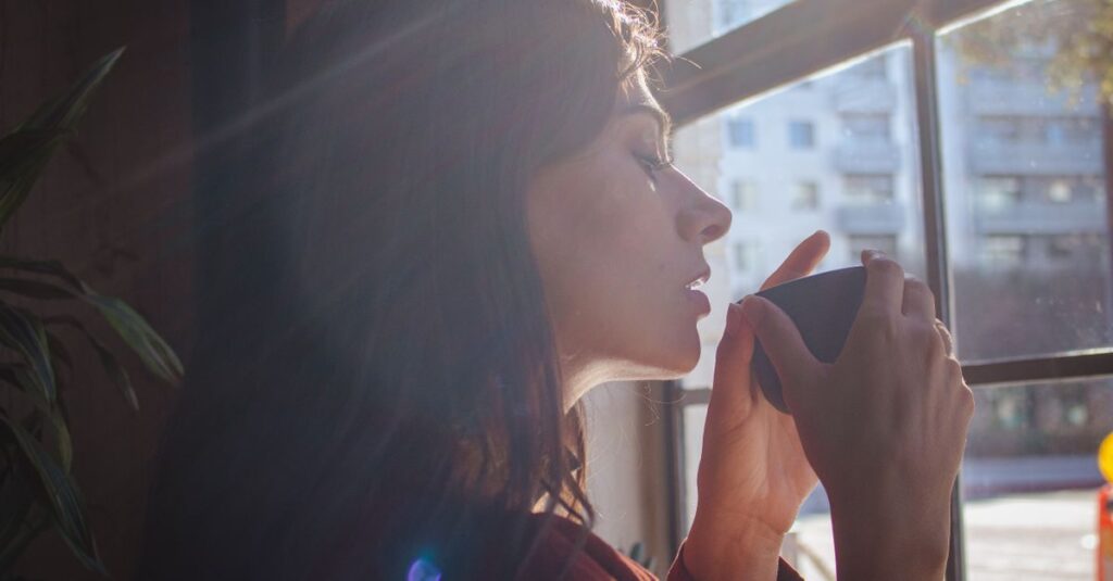 A woman savors a hot drink by a sunlit window in an urban setting.