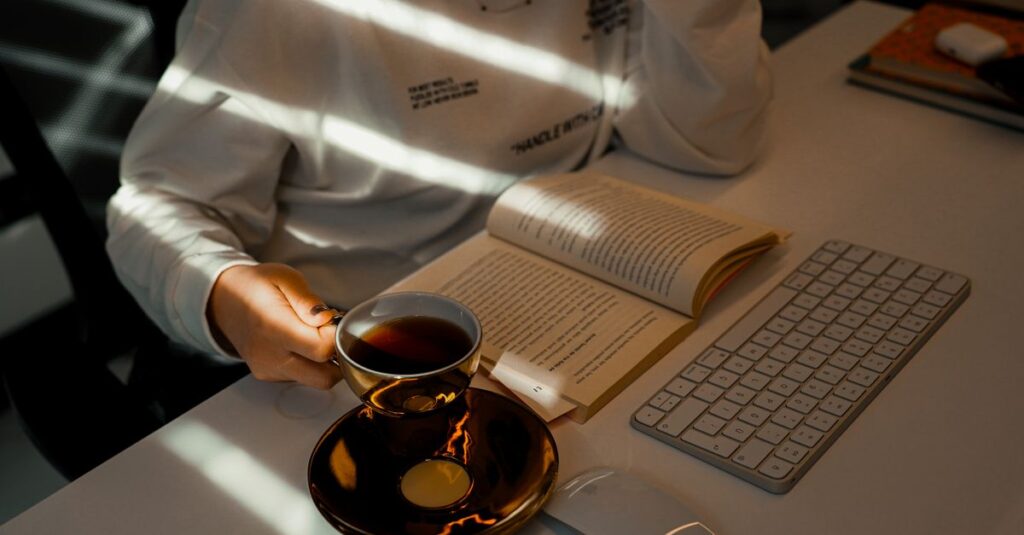 A woman reading a book while holding a cup of tea at a desk with sunlight streaming in.
