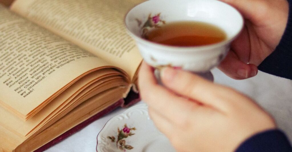 Hands hold a porcelain cup of tea next to an open book, creating a cozy setting.