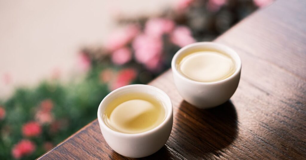Two white tea cups on a wooden table with blurred flowers in the background.