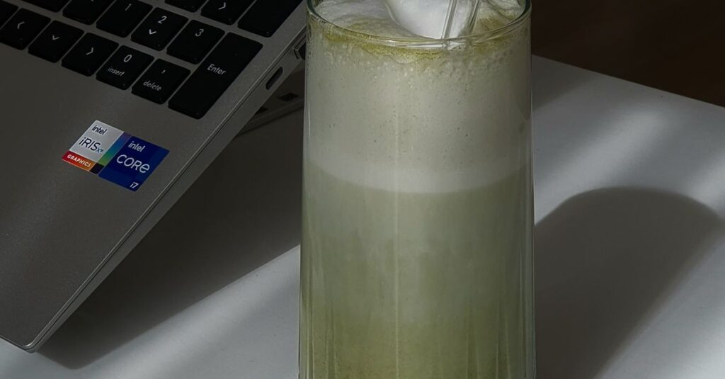 A refreshing iced matcha latte with glass straw on a white desk next to a laptop.