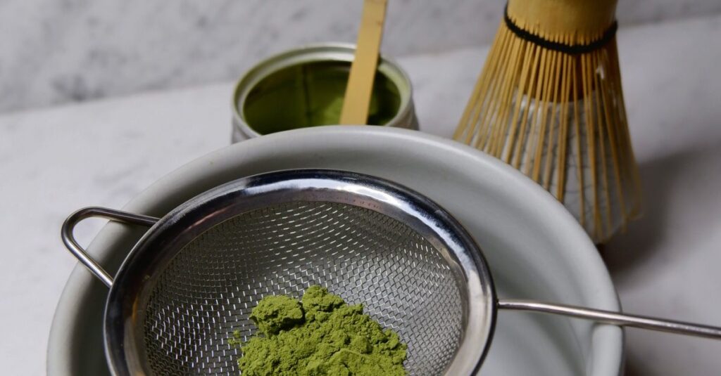 Matcha tea being sifted with a strainer over a bowl, featuring a bamboo whisk and scoop on a marble table.