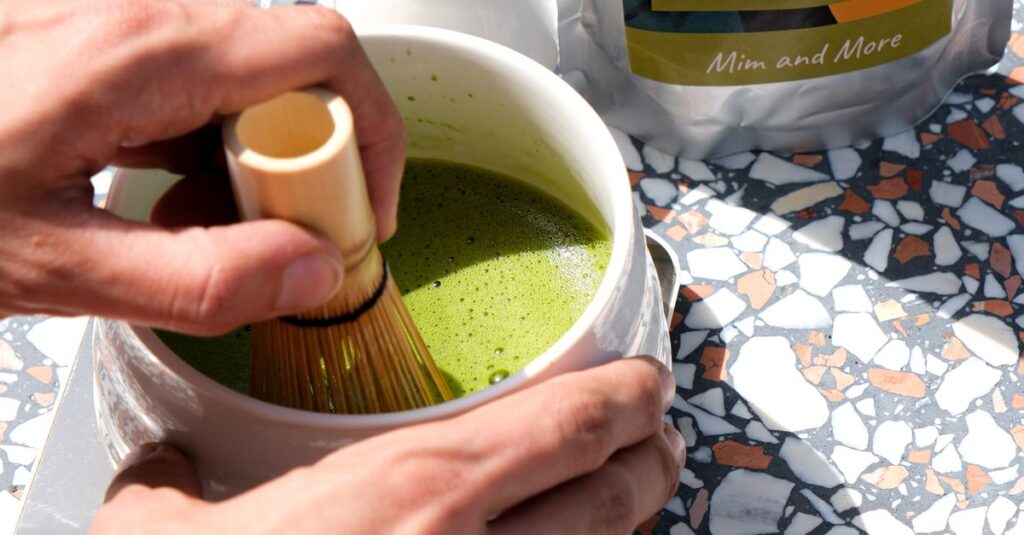 A person whisking vibrant green matcha tea in white bowl using a bamboo whisk on a sunny terrazzo table.