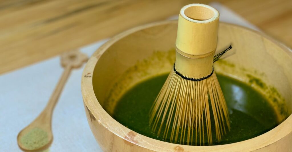 A bamboo whisk in a wooden bowl with matcha tea on a wooden surface.