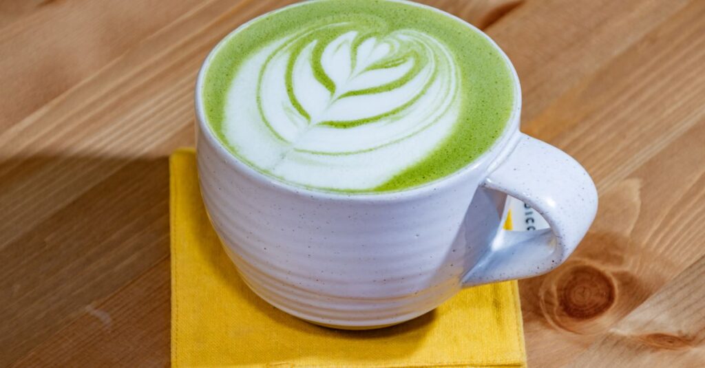 A top view of a green matcha latte with beautiful leaf latte art served in a white cup on a wooden table.