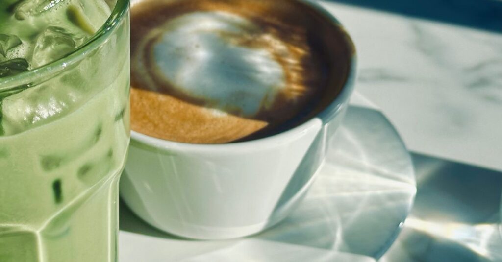 A refreshing iced matcha and cappuccino cup on a sunny marble table, casting artistic shadows.