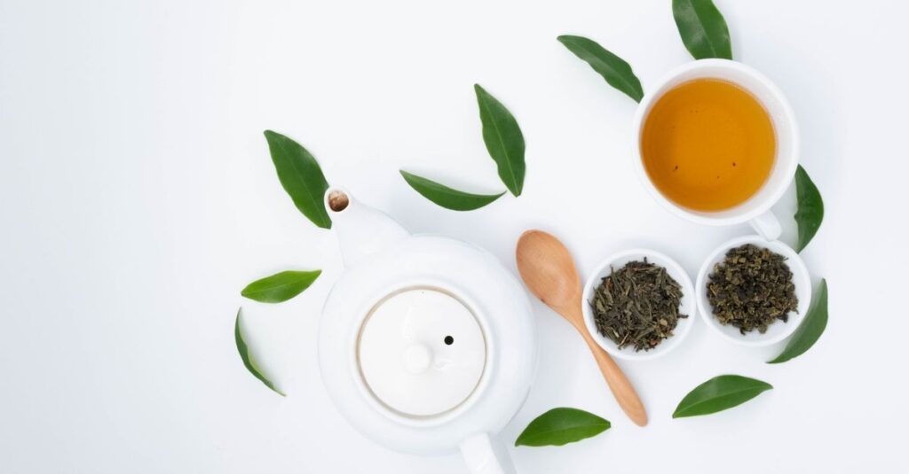 From above of cup with green tea near dry and fresh leaves with ceramic teapot on white table