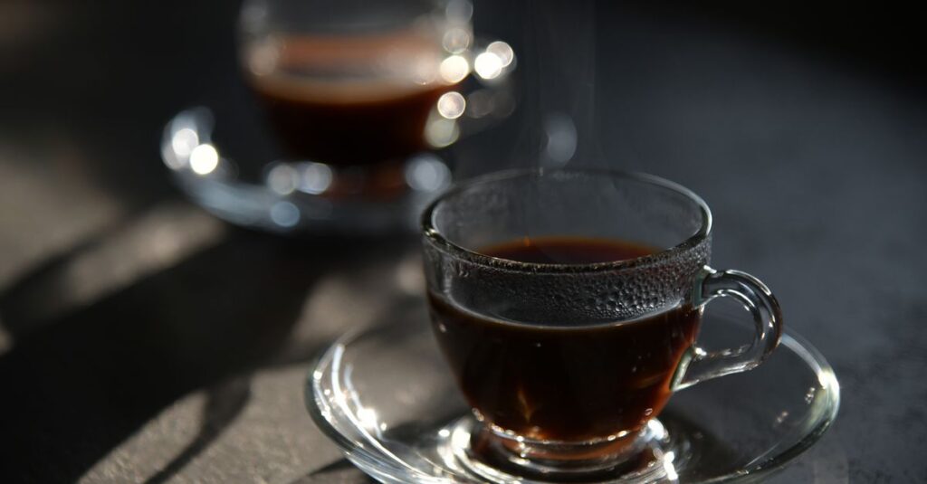 Close-up of steaming black coffee in elegant glass cup with saucer, captured in soft light.