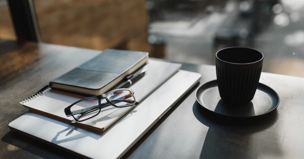 A minimalist workspace with eyeglasses, notebook, and a black coffee cup on a sunlit table.