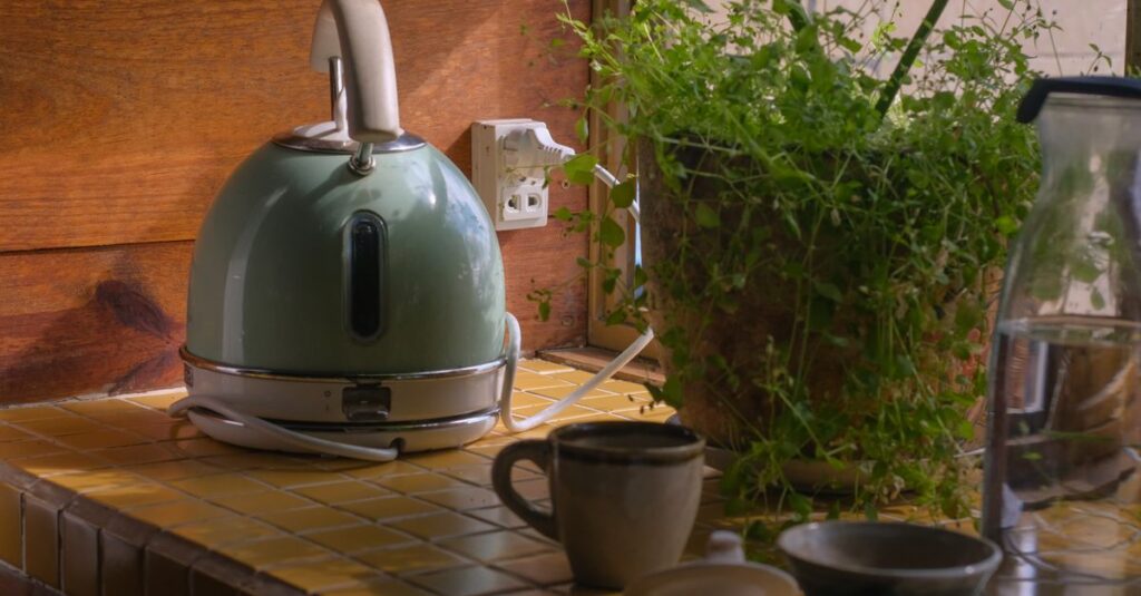 A warm kitchen setup with an electric kettle, potted plant, mugs, and tiled counter.