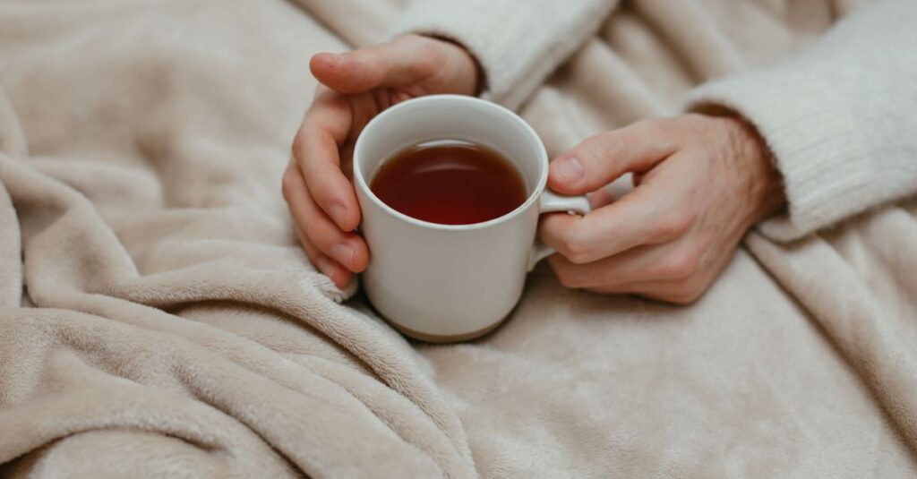 A person holding a ceramic mug filled with tea, wrapped in a warm beige blanket.