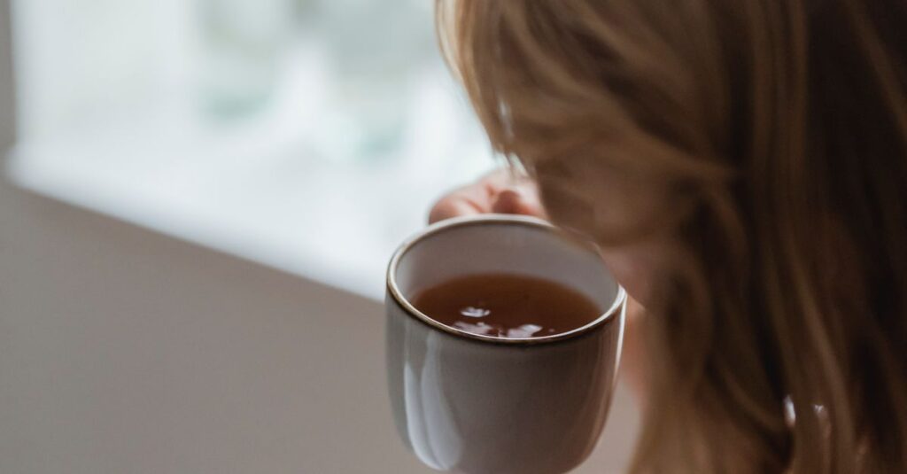 Side view of unrecognizable female with dyed hair in casual clothes drinking aromatic tea while resting in armchair near window at home