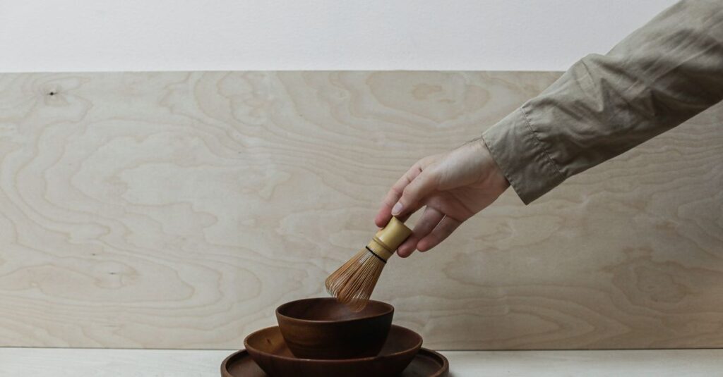 A hand holds a bamboo whisk over stacked wooden bowls, set against a minimalist background.