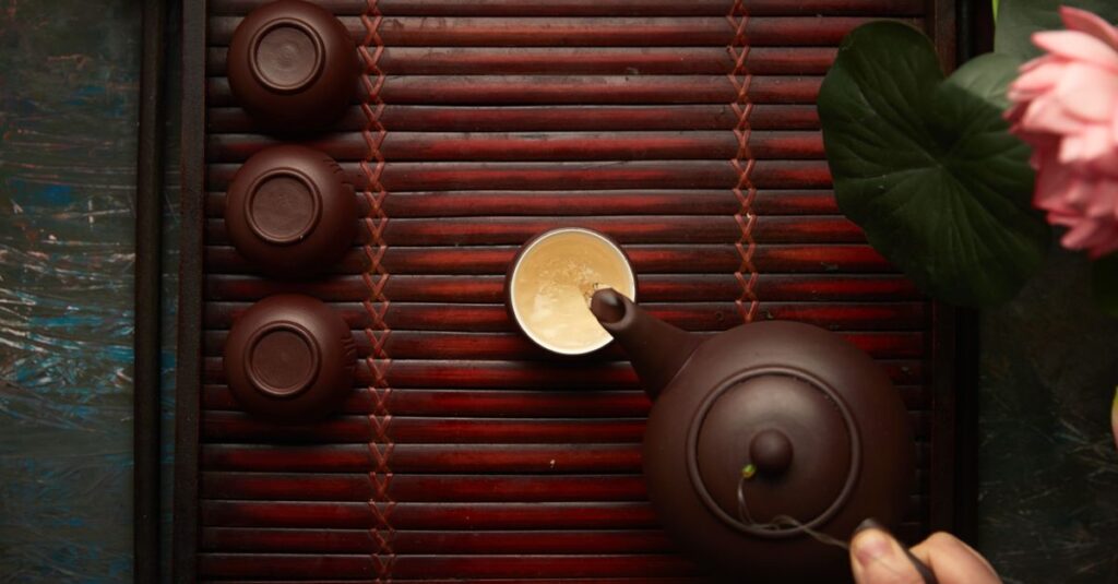 Top view of a hand pouring tea onto a wooden tray with cups, capturing the essence of an elegant tea ceremony.