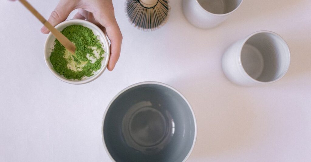Close-up of hands preparing matcha tea with ceramic bowls and bamboo whisk on a white background.