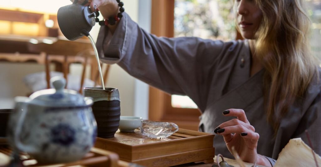 A woman elegantly pours tea from a pot into a cup on a wooden tray, creating a serene setting.