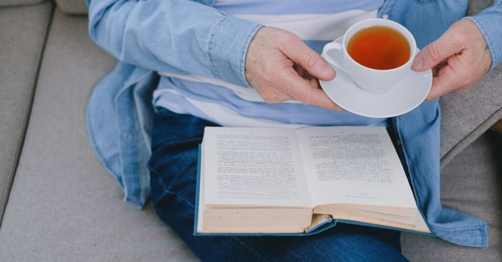 An adult reading a book while holding a cup of tea, seated comfortably indoors.