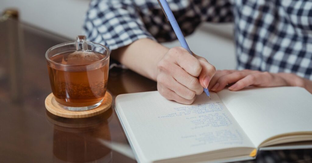 Close-up of hands writing in a notebook beside a glass of tea on a wooden coaster.