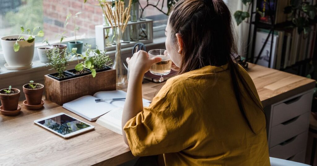 A woman sits at a desk drinking tea, surrounded by plants in a sunlit home office.