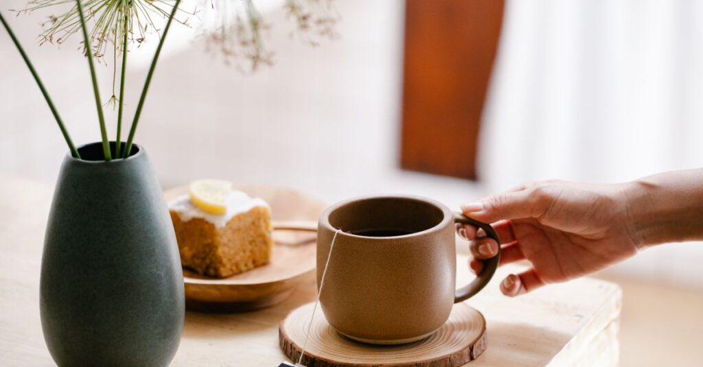 A serene indoor scene with a hand reaching for a mug of tea beside lemon cake and vase.
