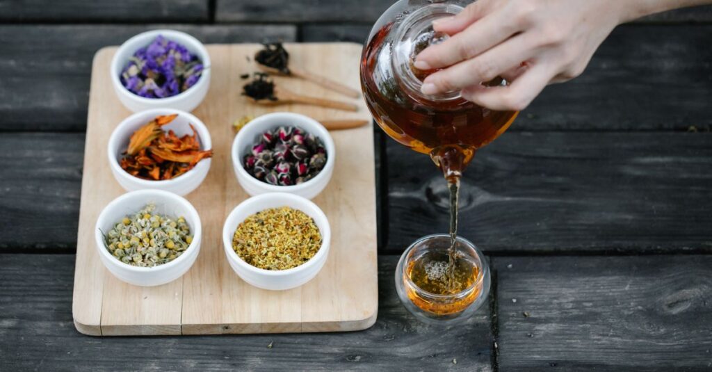 A hand pouring herbal tea into a cup, surrounded by various dried herbs on a cutting board.