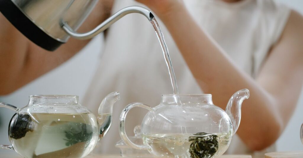 A woman pours hot water into glass teapots with blooming tea indoors.