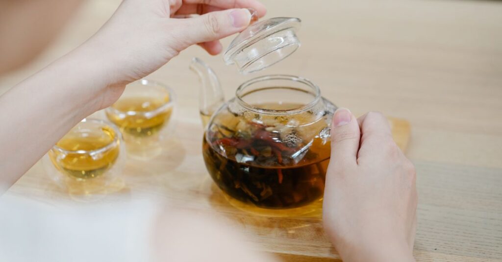 Hands lifting the lid of a glass teapot with herbal tea and two teacups nearby.