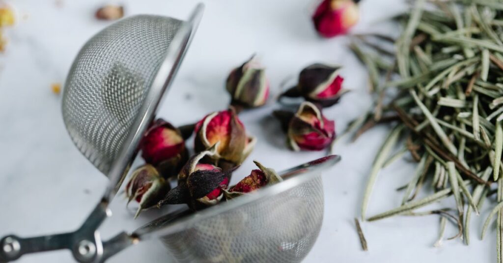 Close-up of rosebuds and herbs in a stainless steel tea strainer on a white surface.