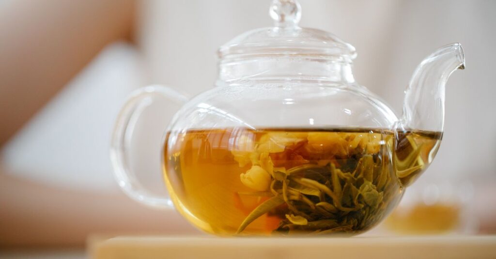 A clear glass teapot containing a brewed herbal tea blend placed on a wooden surface indoors.