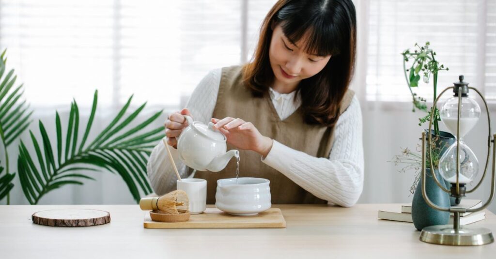 Asian woman using a teapot and bamboo whisk to pour tea indoors at a cozy table setting.